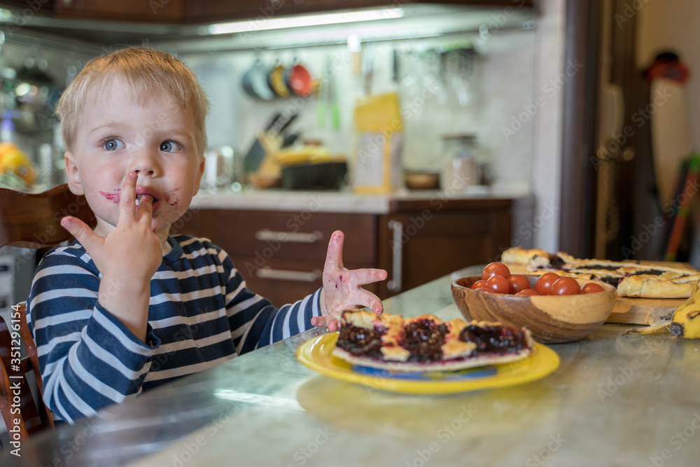 Little cute blond boy licks sweet fingers while eating delicious homemade berry pie in kitchen. Tasty food for kids Little cute blond boy licks sweet fingers while eating delicious homemade berry pie in kitchen. Tasty food for kids