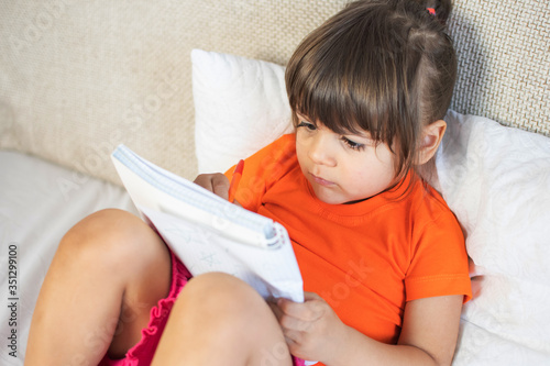 Little brown hair girl with brown eyes. 3 years old. Painting with pen in notebook. Sitting on sofa at home.