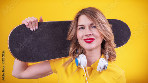 Happy lady in casual yellow polo with red lips and headphones holding skateboard on shoulder and looking at camera while standing on yellow background
