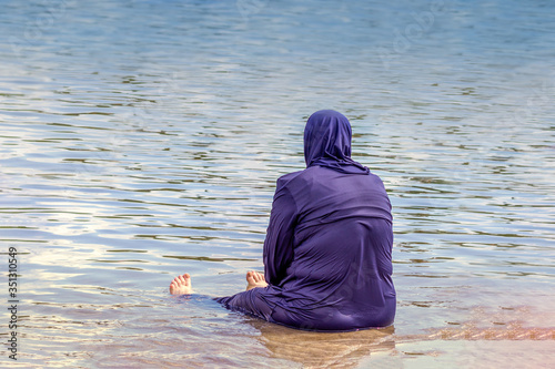 A Muslim woman dressed in a national costume for swimming sits on the seashore and looks into the distance. View from the back