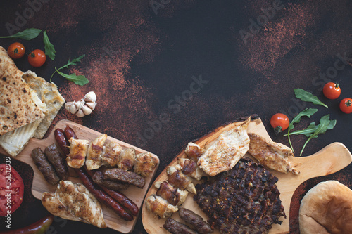 Various grilled meats served on wooden cutting boards with fresh vegetables and bread on a dark rustic background. Traditional Serbian and Balkan bbq meat called rostilj. Flat lay. Copy space photo