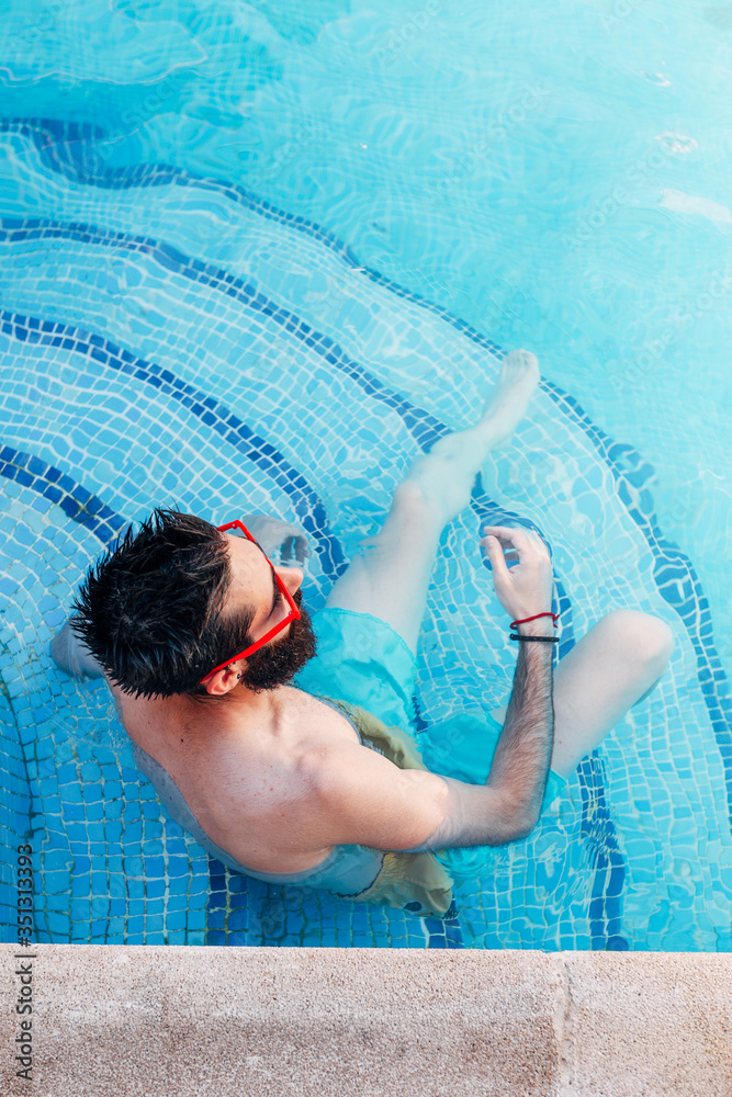 Top view of sexy young man with beard and red sunglasses sitting on the pool steps. Vacation concept.