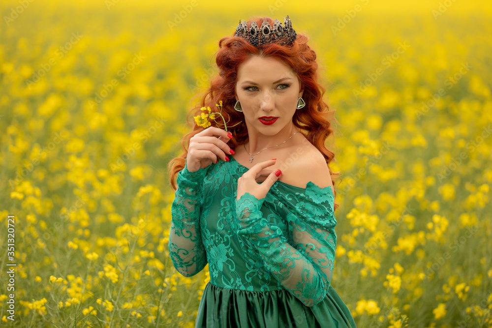 Beautiful Redhead female model in a field of yellow rapeseed flowers. Wearinf at long green dress and crown Beautiful Redhead female model in a field of yellow rapeseed flowers. Wearinf at long green dress and crown