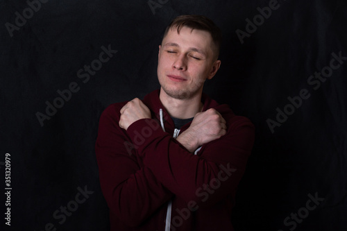 Studio portrait of a young man in a red sweatshirt against black background. A deaf-mute guy shows a "love" gesture with his fingers. Good eyes. Facial expression and sweet smile. Sign language. © Anton