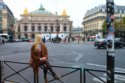 Blonde girl in a brown coat in front of the Palace or Opera Garnier and the National Academy of Music. Automobiles and pedestrians in front of the Grand Opera.