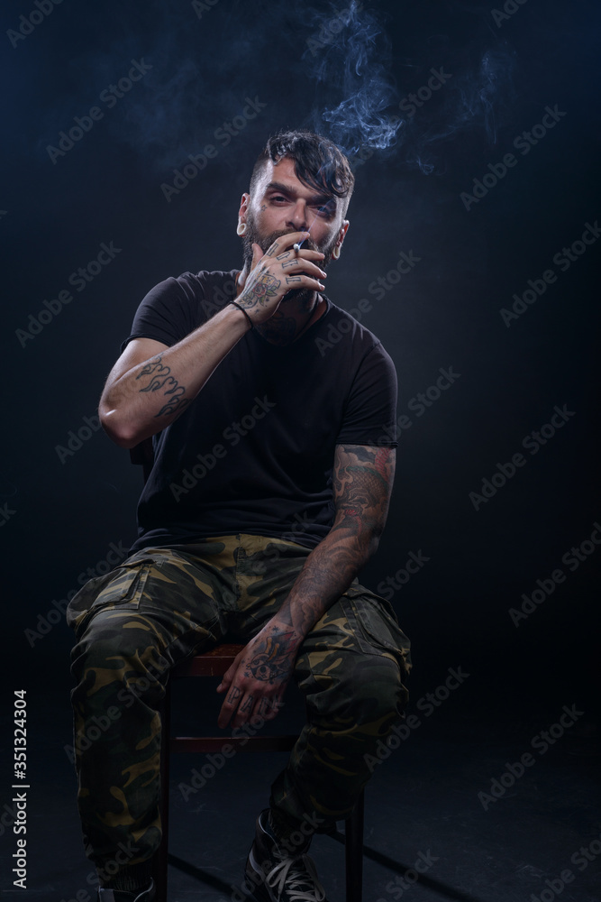 Portrait of a handsome young tattooed man smoking a cigarette isolated in a studio on a dark background. Portrait of a handsome young tattooed man smoking a cigarette isolated in a studio on a dark background.
