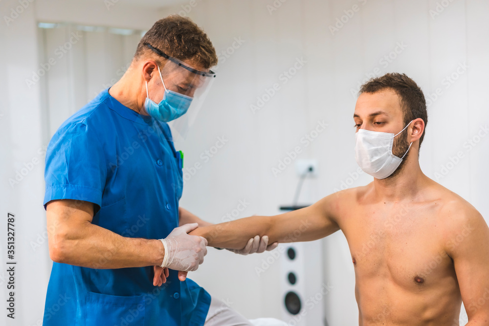 Obraz premium A physiotherapist with a screen and mask giving a wrist massage to a patient with a mask. Physiotherapy with protective measures for the Coronavirus pandemic, COVID-19. Osteopathy, therapeutic chiroma
