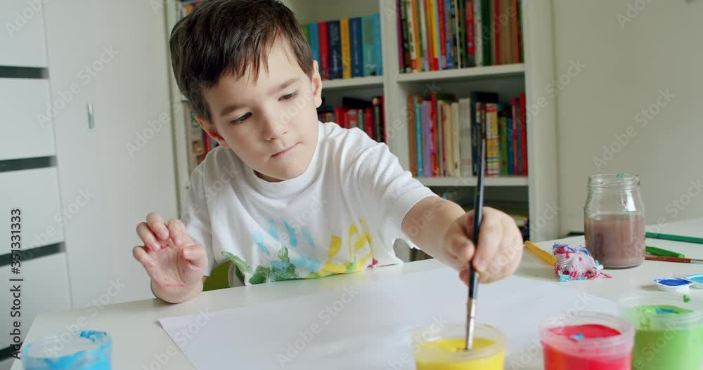 School boy reaching for paints with paintbrush in his hand and painting. Child with paintbrush and colorful paints at home. Hand-held shot, single shot, medium close up, 4K. School boy reaching for paints with paintbrush in his hand and painting. Child with paintbrush and colorful paints at home. Hand-held shot, single shot, medium close up, 4K.
