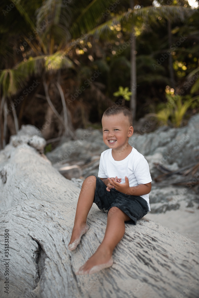 Happy smile european blond baby boy have rest and sit on white sand beach. . white cotton clothes. t-short.Thailand. palms. rocks Happy smile european blond baby boy have rest and sit on white sand beach. . white cotton clothes. t-short.Thailand. palms. rocks