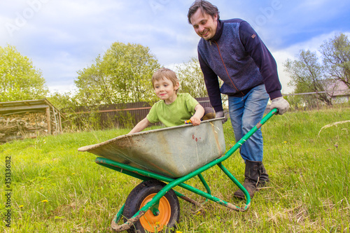 Man rolls his son in a garden cart in the garden. The son laughs and rejoices.