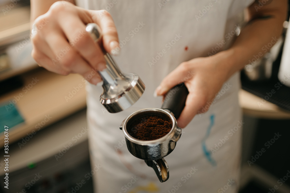 A close-up photo of female hands holding a metal tamper and a portafilter with coffee in a coffee shop. A barista preparing for pressing ground coffee for brewing espresso or americano in a cafe.