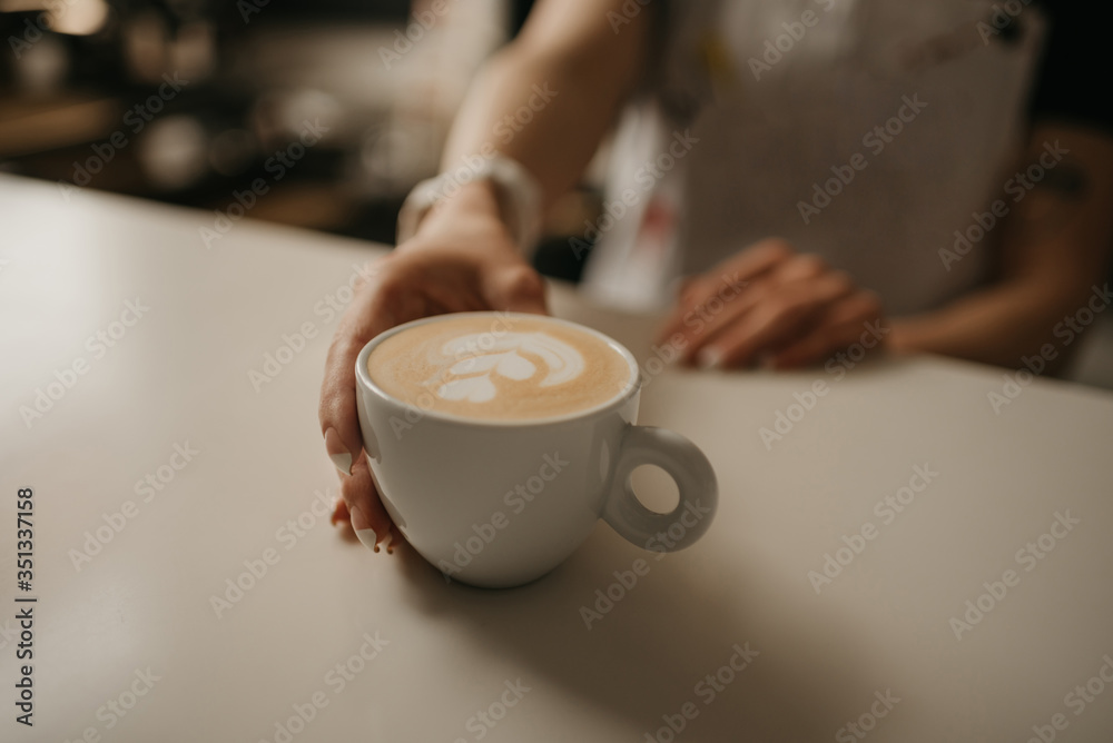A female barista holds out a cup of hot latte to a client in a cafe. A waitress preparing a customer order.
A female barista holds out a cup of hot latte to a client in a cafe. A waitress preparing a customer order.