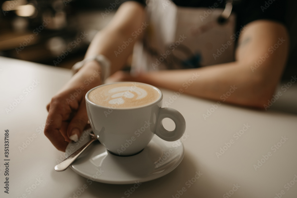 A female barista holds out a cup of hot latte with a saucer and spoon to a client in a cafe. A barista preparing a customer order. A female barista holds out a cup of hot latte with a saucer and spoon to a client in a cafe. A barista preparing a customer order.