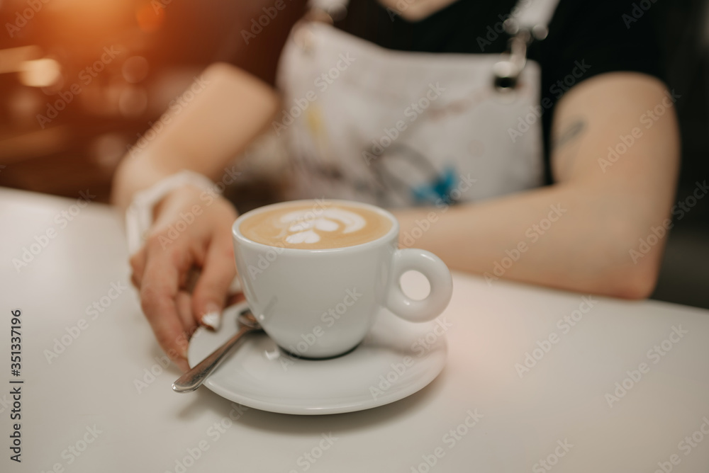 A female barista holds out a cup of fresh latte with a saucer and spoon to a client in a cafe. A barista preparing a customer order. A female barista holds out a cup of fresh latte with a saucer and spoon to a client in a cafe. A barista preparing a customer order.