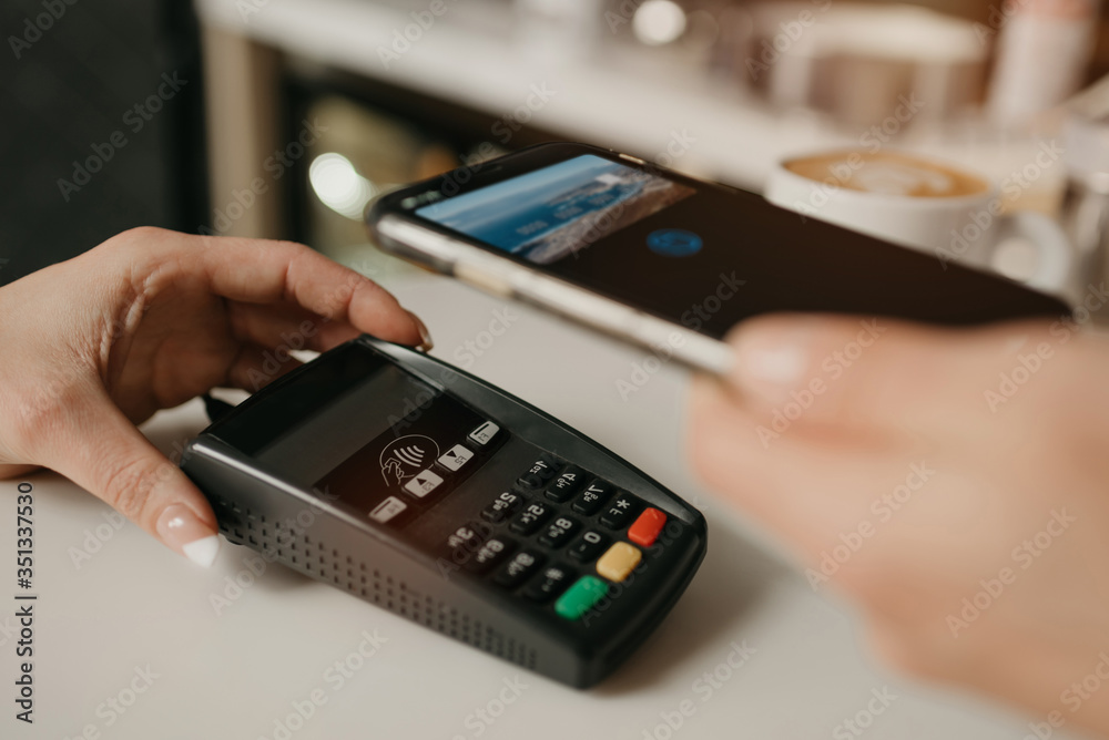 A lady paying for her latte with a smartphone by contactless NFC technology in a cafe. A female barista holds out a terminal for paying to a client in a coffee shop. A lady paying for her latte with a smartphone by contactless NFC technology in a cafe. A female barista holds out a terminal for paying to a client in a coffee shop.