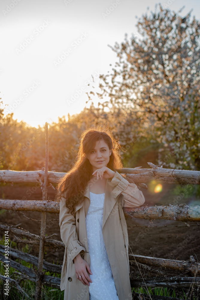Naklejka premium A young woman in a white dress and beige trench coat stands near a wooden fence in nature, a beautiful girl with long hair in the countryside