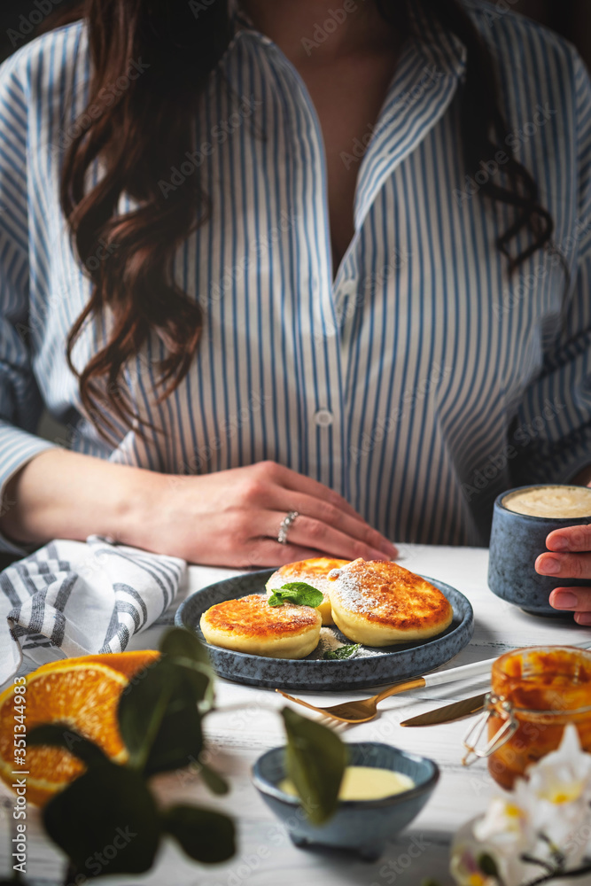 Woman at the table with a beautiful and tasty breakfast. Cottage cheese pancakes on a plate, cup with coffee and oranges on a white wooden background.