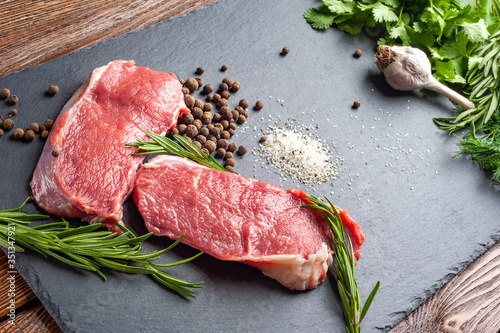 Raw beef meat and spices. Slices of fresh raw meat, rosemary and fresh herbs on a black slate stone plate. Preparing and marinating barbecue meat.