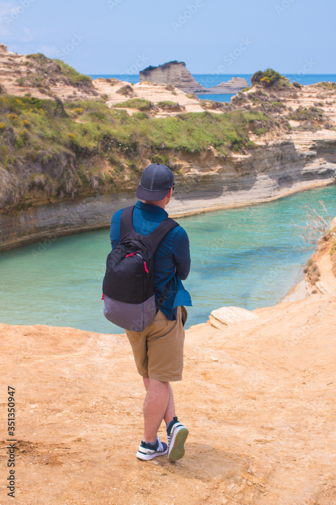 Young man traveler wearing stylish casual cloth,cap,backpack standing on empty edge of high sandy cliff rock on blue sea background.View from the back. Young student male hipster exploring.