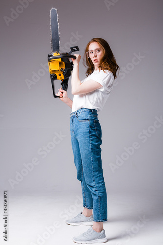 Attractive redhead girl in a white T-shirt and jeans holds up a chainsaw vertically and looks at the camera on a gray background