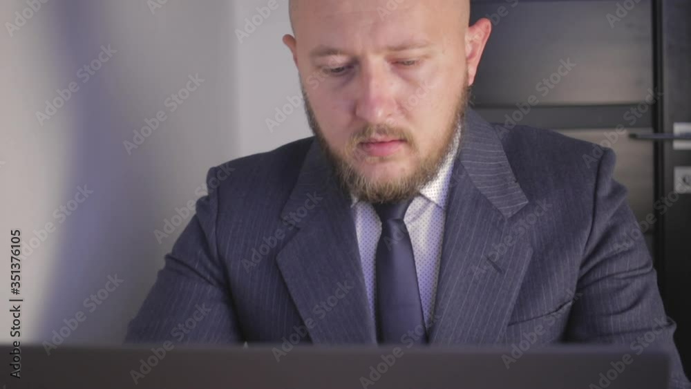 Caucasian bald young man with beard in suit works on laptop and looked in camera, close-up shot Caucasian bald young man with beard in suit works on laptop and looked in camera, close-up shot