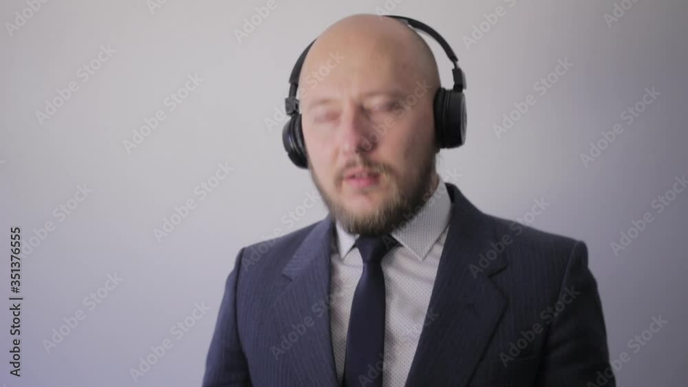 Bald young man with beard in suit looking in camera, uses wireless headphone and listen music, close-up view Bald young man with beard in suit looking in camera, uses wireless headphone and listen music, close-up view