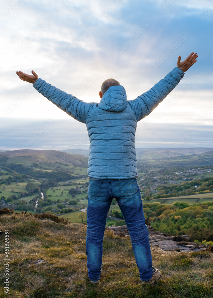 Man wearing grey coat and jeans raising his hands high and standing on top of mountain   