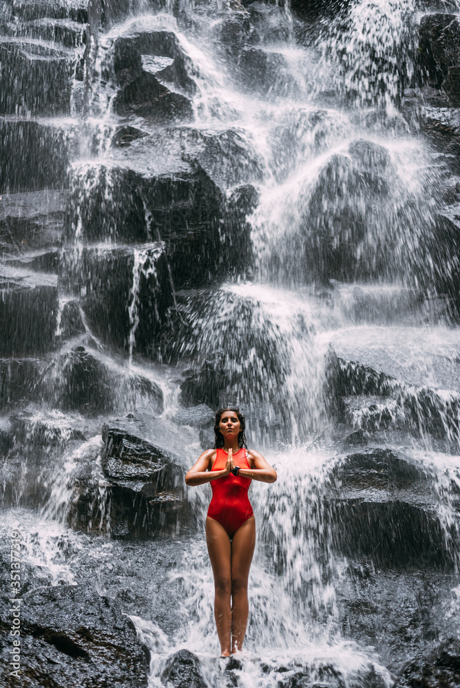 Obraz premium A woman does yoga at a waterfall. Healthy lifestyle. The concentration of the body. A woman does yoga in Bali. A woman meditates in nature. Meditation at the waterfall. Young woman practicing yoga