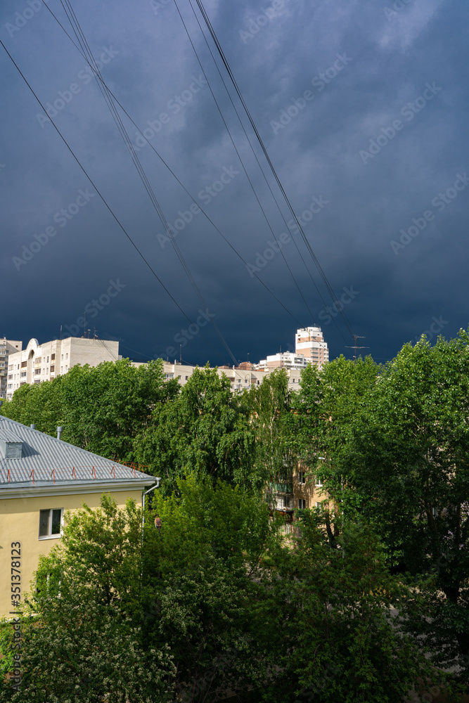 Town landscape before the storm with sunlit buildings, green trees and dark gloomy sky