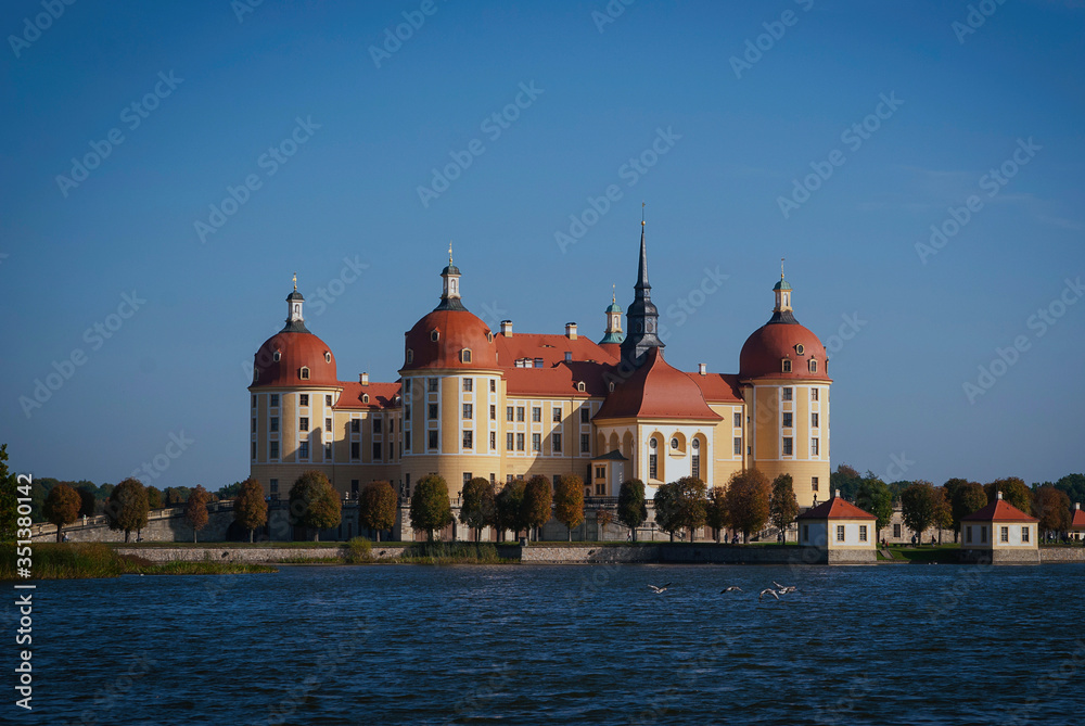 View of Moritzburg Castle, Germany from a distance. Beautiful yellow castle with a red roof against a blue sky on the river bank on a sunny day.