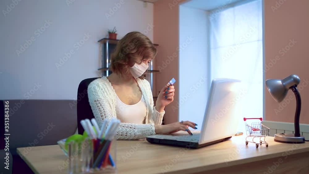 The girl works at home during quarantine. Coronavirus pandemic. A girl in a surgical mask works on a laptop, enters data from a card. Remote work. The girl works at home during quarantine. Coronavirus pandemic. A girl in a surgical mask works on a laptop, enters data from a card. Remote work.