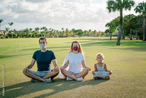 the family in the park practices yoga in protective masks against the coronavirus covid-19. Threesomes are sitting in a clearing on a grass sunny day. lotus position