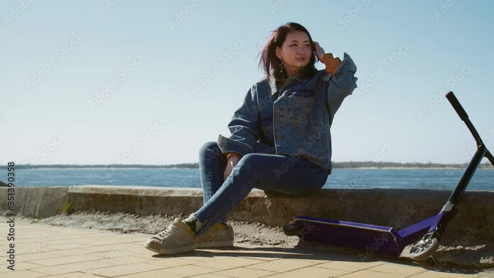 Young asian woman in a jeans jacket sits on the border with the scooter near the river Young asian woman in a jeans jacket sits on the border with the scooter near the river