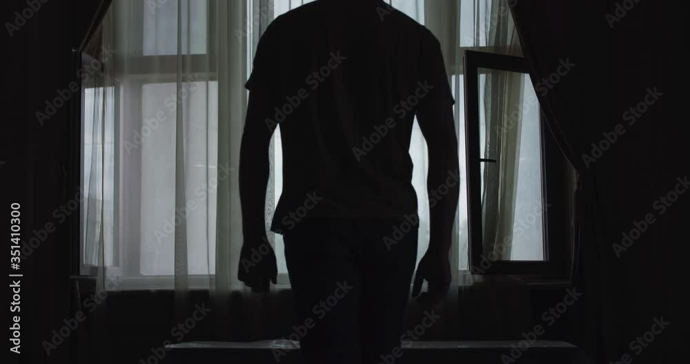 Silhouette of man comes to light window in dark room and crosses hands Silhouette of man comes to light window in dark room and crosses hands