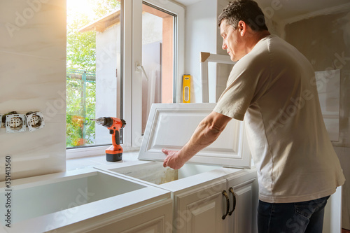 Middle-aged Caucasian man holds kitchen cabinet door , preparing for the process of custom installation in his own house. Quarantine activity, repair concept