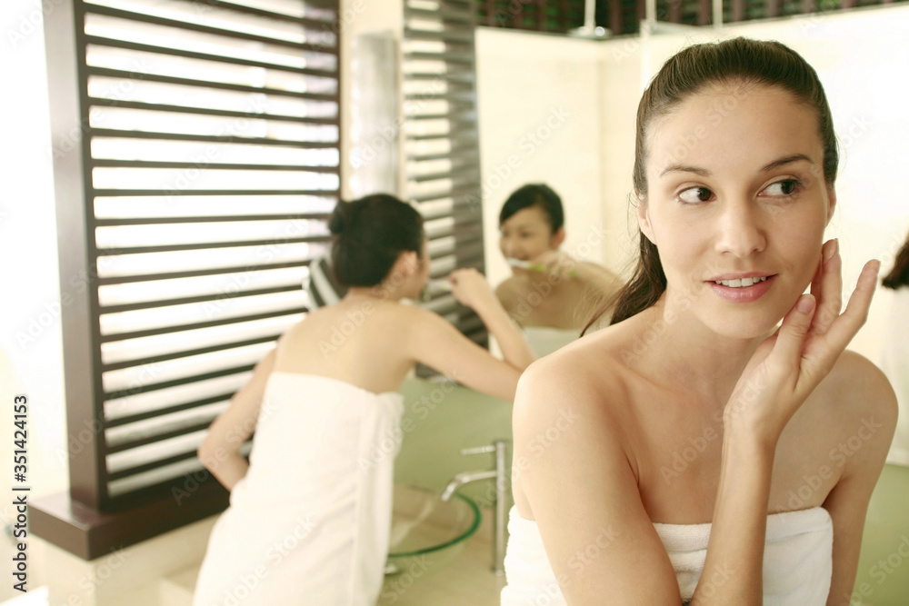 Woman applying moisturizer on her face, friend brushing teeth in the background Woman applying moisturizer on her face, friend brushing teeth in the background