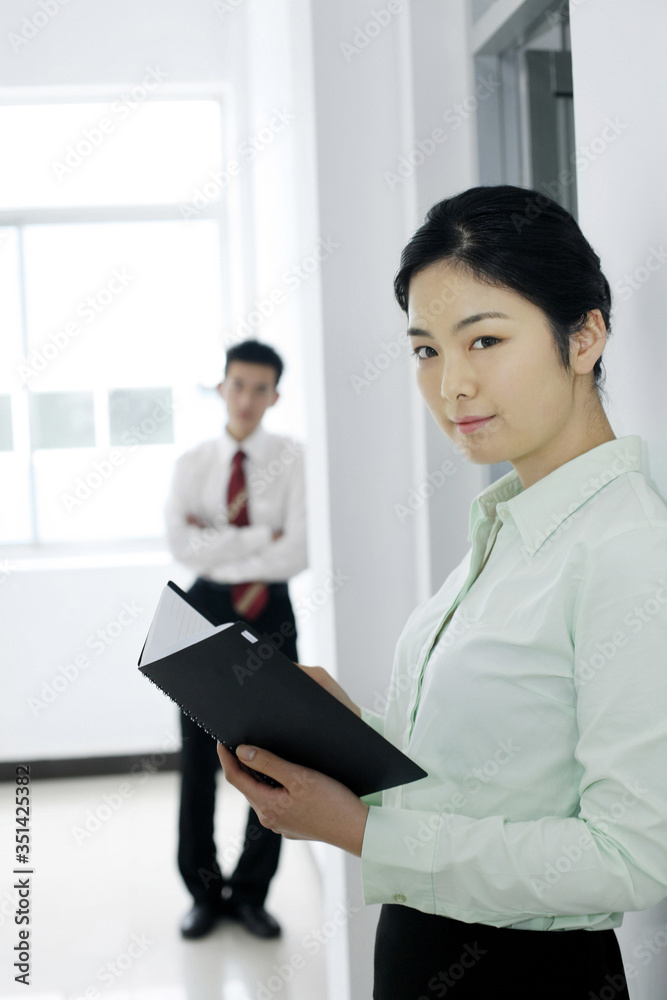 Businesswoman holding organizer with businessman standing in the background Businesswoman holding organizer with businessman standing in the background