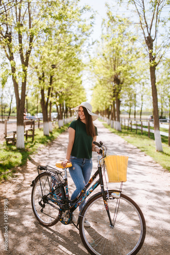 A beautiful young woman with a straw hat, bicycle and flowers is standing in the park. Life in a village