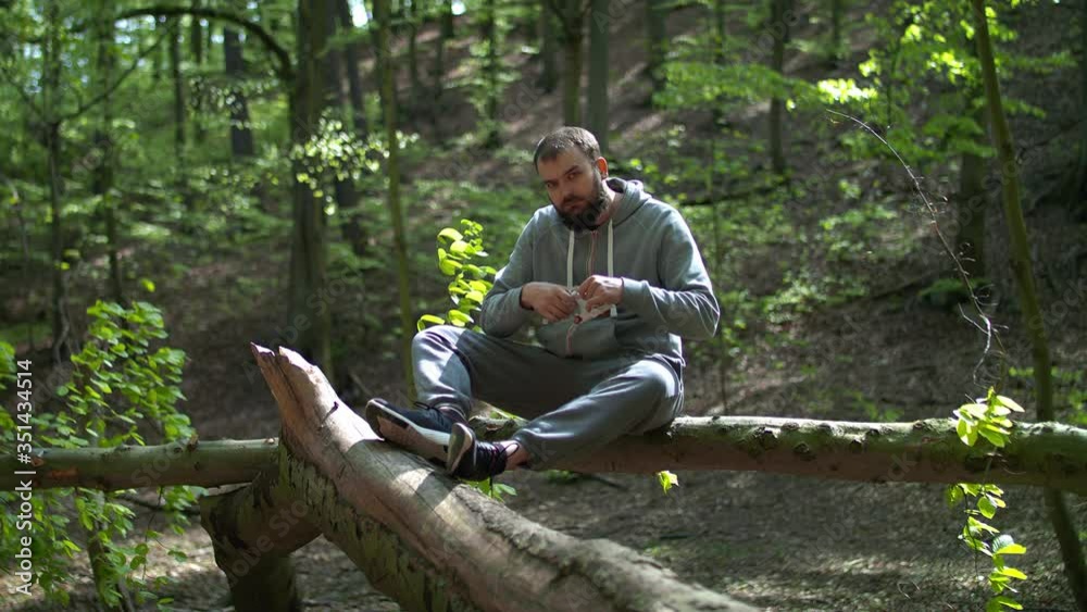 Young Bearded Man Sitting on Tree Branch and Putting on Protective Face Mask