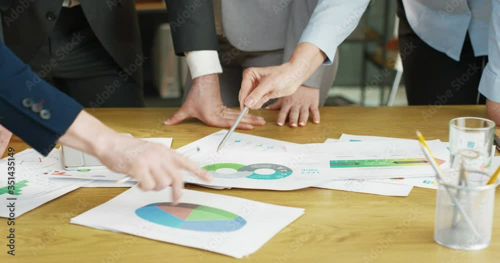 Close up of papers and documents with charts, graphics and diagrams lying on wooden table in office. Business meeting and discussion concept. Mixed-races female and male hands. Multiethnic partners. Close up of papers and documents with charts, graphics and diagrams lying on wooden table in office. Business meeting and discussion concept. Mixed-races female and male hands. Multiethnic partners.