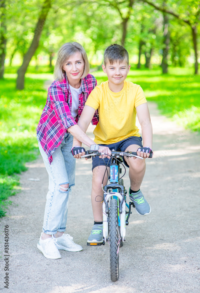 Sporty family leisure. Woman teaches her young son ride a bike in summer park. Empty space for text