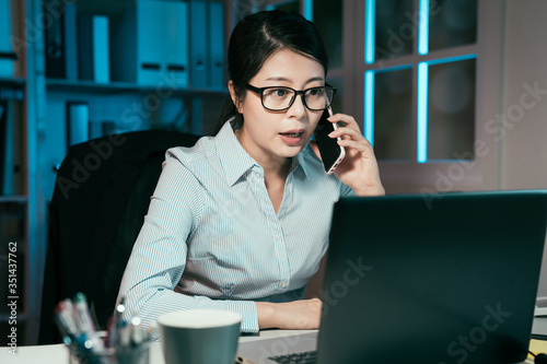 business overwork deadline and people concept. happy asian korean businesswoman worker with laptop computer calling on smartphone at night office. lady having discussion on new plan with coworker.