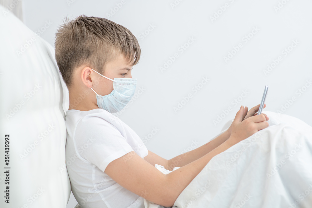 Sick young boy wearing medical protective mask uses tablet computer at home. Quarantine and coronavirus epidemic concept Sick young boy wearing medical protective mask uses tablet computer at home. Quarantine and coronavirus epidemic concept
