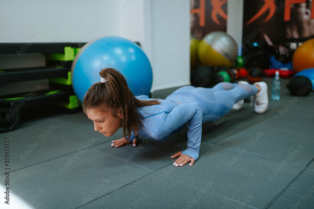 Fototapeta premium A beautiful young caucasian girl in blue sports equipment does push-ups in the gym