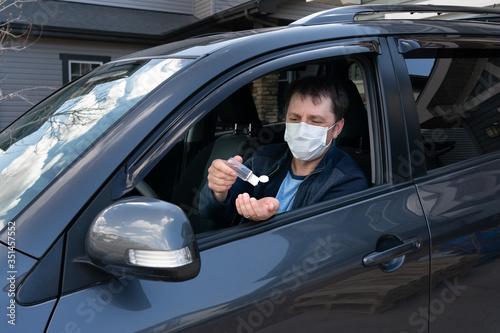 Middle age man in protective sterile medical mask driving car going to buy groceries.The concept of preventing the spread of coronavirus.A man wearing mask sits in dark grey car uses hand sanitizer. © Oksana