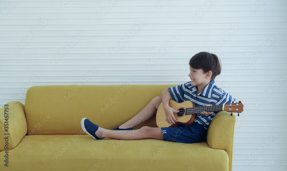 Portrait side of little Caucasian boy sitting on yellow sofa and playing ukulele guitar in living room at home Portrait side of little Caucasian boy sitting on yellow sofa and playing ukulele guitar in living room at home