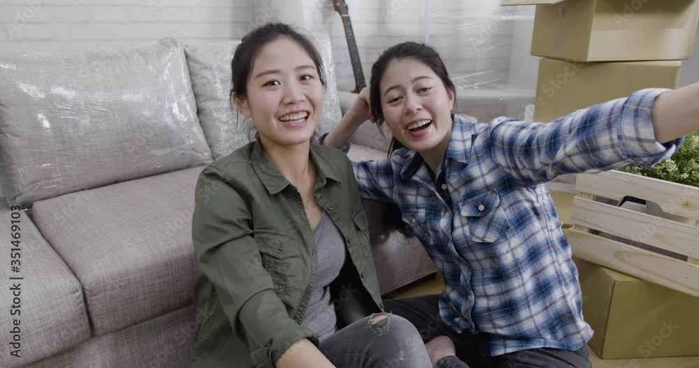 two young asian women moving into new home talking on mobile phone with video face camera showing house apartment. sisters chatting with parents in living room sit floor. people technology relocation two young asian women moving into new home talking on mobile phone with video face camera showing house apartment. sisters chatting with parents in living room sit floor. people technology relocation