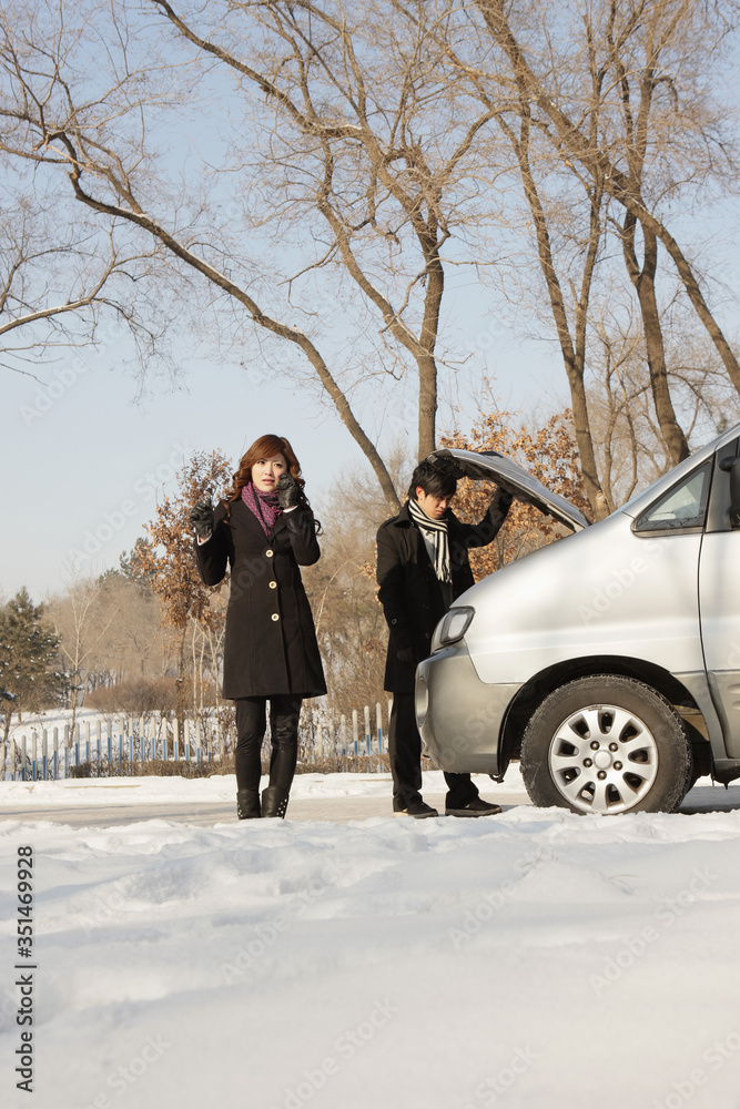 Businessman looking under hood of car, businesswoman talking on the mobile phone