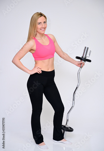 A slender Caucasian model poses in various poses with a smile. Portrait of a pretty fitness blonde girl in a sports uniform on a white background in full length with a barbell in her hands.