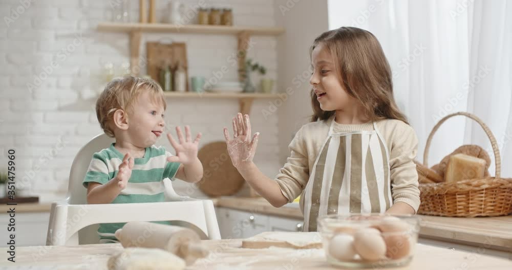 Little girl is preparing cake dough while her brother plays in baby chair, then sister does highfive with floured hand, boy happily laughing - togetherness 4k Little girl is preparing cake dough while her brother plays in baby chair, then sister does highfive with floured hand, boy happily laughing - togetherness 4k